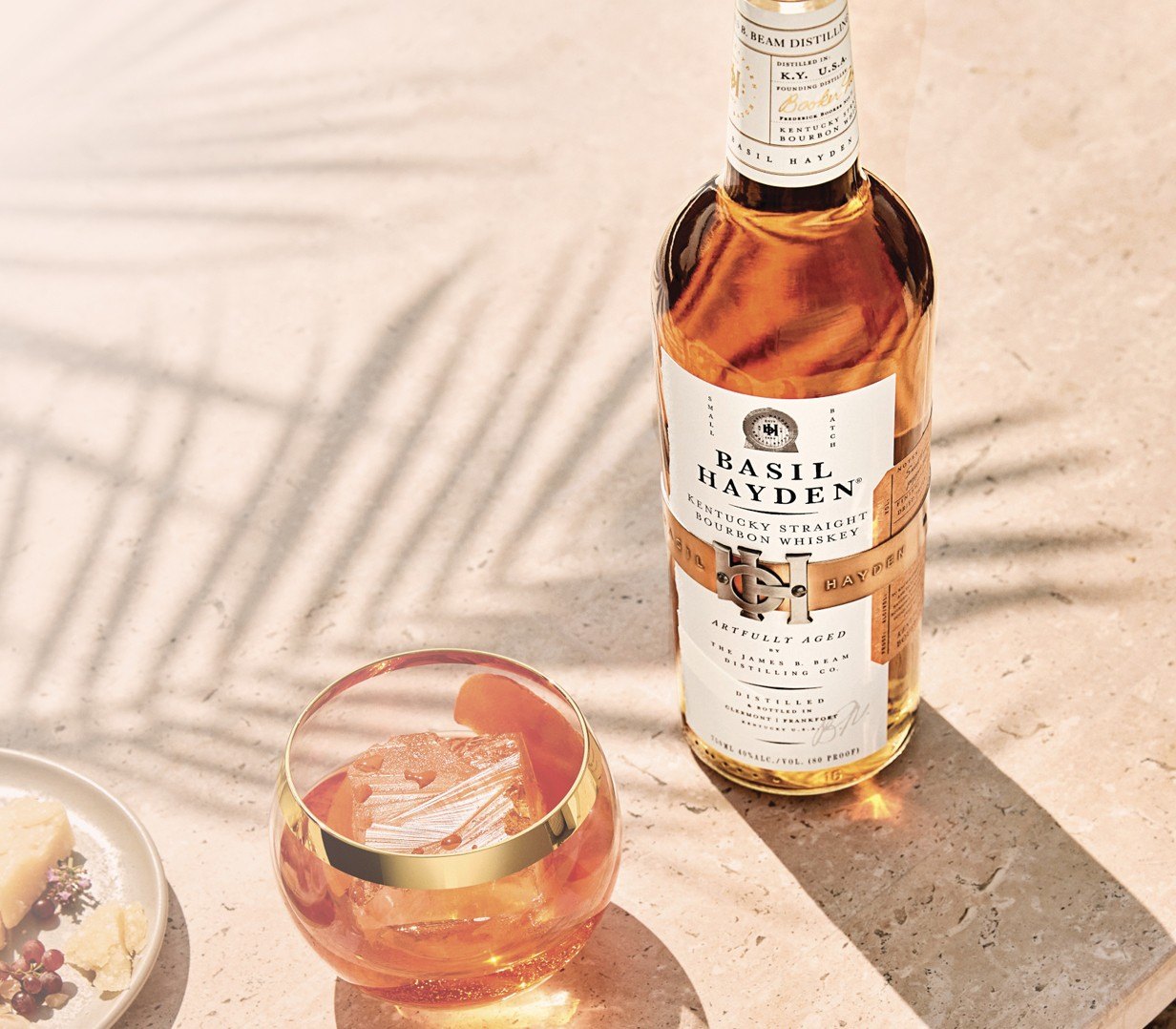 A bottle of Basil Hayden bourbon beside a cocktail over ice on a sunlit stone table.